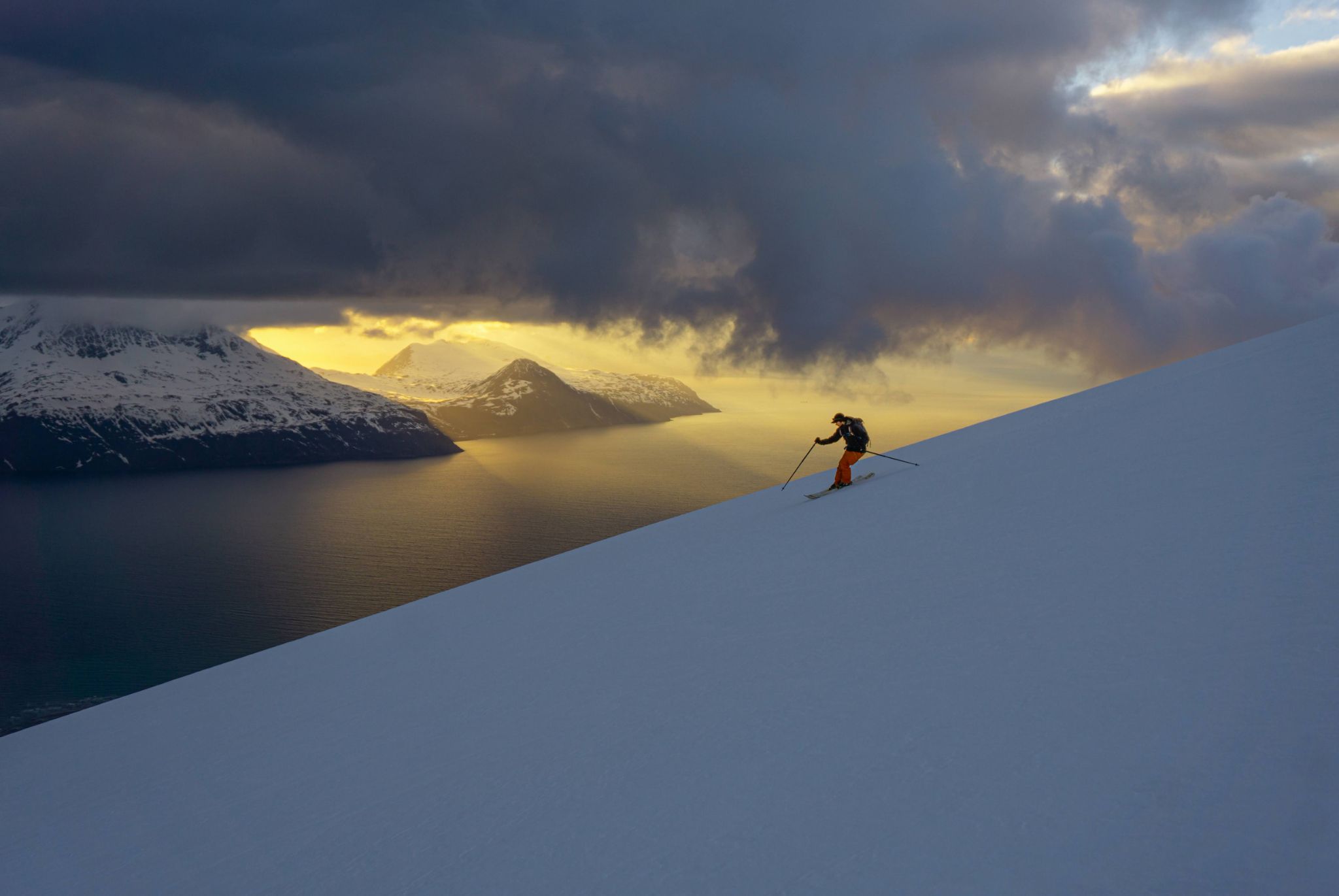 Skiers climbing a snow ridge above a fjord under bright clear skies